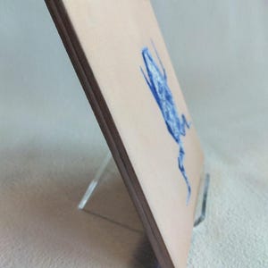 May include: A square, light-tan artwork with a blue illustration of a creature, possibly a frog or insect. The artwork is displayed on a clear acrylic stand. The edges of the artwork are dark brown.