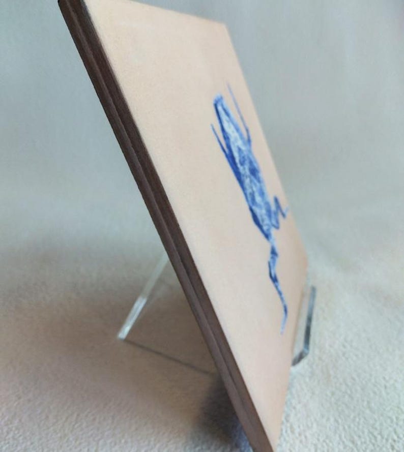 May include: A square, light-tan artwork with a blue illustration of a creature, possibly a frog or insect. The artwork is displayed on a clear acrylic stand. The edges of the artwork are dark brown.