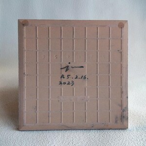 May include: A square, light brown ceramic tile with a grid pattern on the back. The tile has a handwritten signature and the date "R 5.2.16. 2023" in black ink. The edges of the tile are slightly darker.