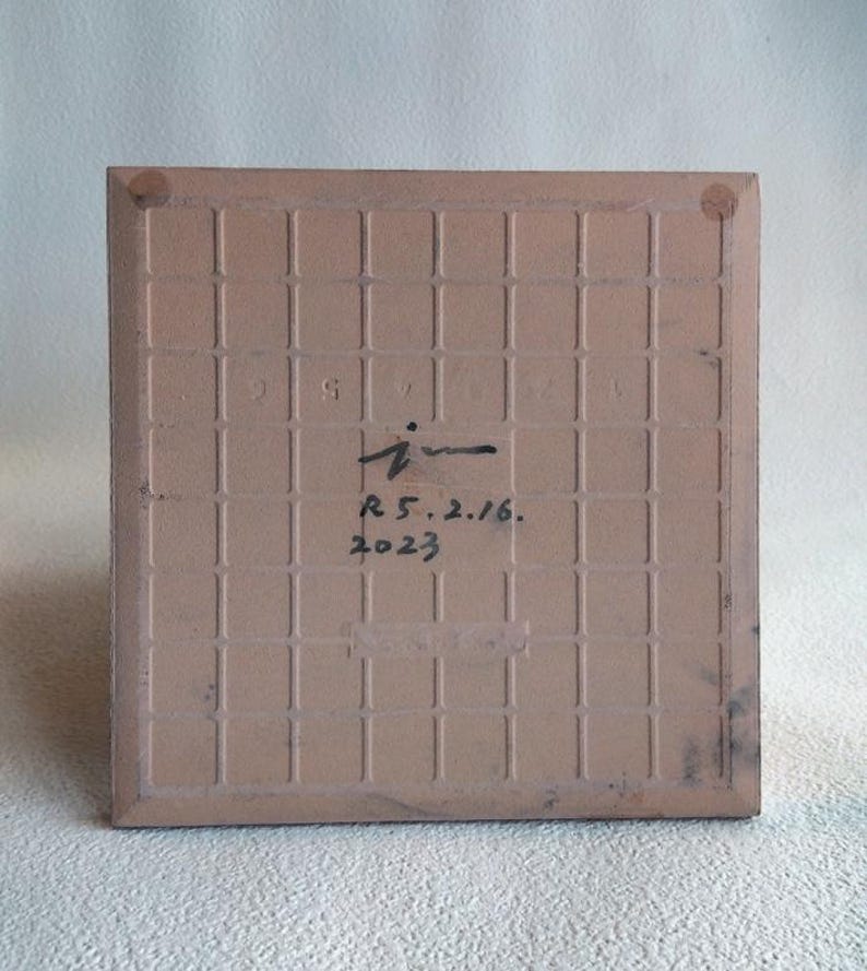 May include: A square, light brown ceramic tile with a grid pattern on the back. The tile has a handwritten signature and the date "R 5.2.16. 2023" in black ink. The edges of the tile are slightly darker.