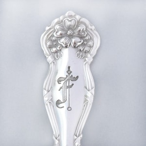 May include: Silverware handle with a floral design and the letter "F" engraved on the front.