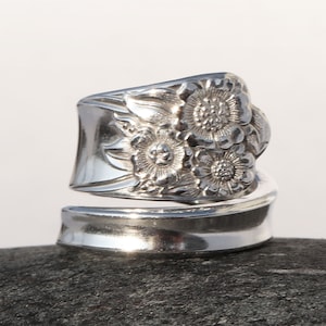 May include: A silver ring made from a repurposed spoon with a floral design. The ring is wide and has a curved shape. It is sitting on a gray rock.