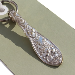 May include: Silver spoon keychain with a floral design. The spoon has a key ring attached to the top and features a detailed floral pattern with two large flowers and smaller floral accents.