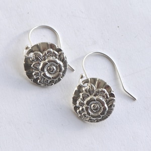 May include: A pair of silver-toned earrings. Each earring features a circular design with a detailed floral pattern. The earrings have a hook closure. The background is a plain white surface.