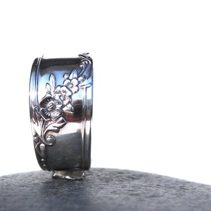 May include: A silver-colored ring with a floral design. The ring features embossed flowers and leaves. The ring is standing upright on a dark gray surface against a white background. The ring is made of metal.