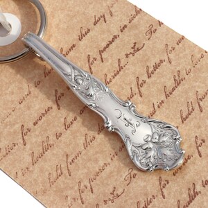 May include: A silver spoon keychain with ornate floral and leaf detailing. The spoon's handle is engraved with initials. The keychain is attached to a white ribbon and a silver ring. The background is a brown paper with handwritten text.