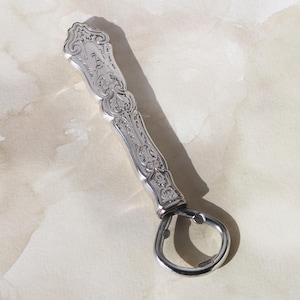 May include: Silver bottle opener with ornate, engraved detailing. The handle features a decorative design with an initial. The opener has a loop for leverage and is likely made of stainless steel. A classic barware accessory.