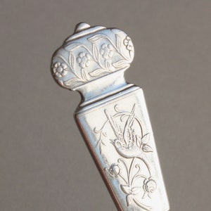 May include: Close-up of a silver utensil with intricate floral and bird engravings. The handle features detailed flower patterns, while the body showcases a bird within a circular frame, surrounded by floral designs. The utensil has a decorative, ornate design.