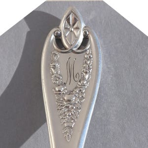 May include: Close-up of a silver spoon with ornate floral and leaf detailing. The spoon features an engraved initial "M" and a decorative design at the handle's end. The spoon is likely vintage or antique, showcasing detailed craftsmanship.