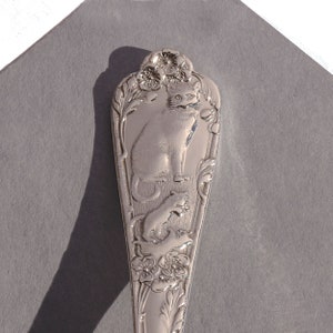May include: A silver spoon with an ornate design featuring a cat and kittens. The spoon is decorated with flowers and leaves.