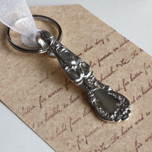 May include: Silver spoon keychain with an ornate design and a key ring attached. The spoon is on a brown paper background with cursive text.
