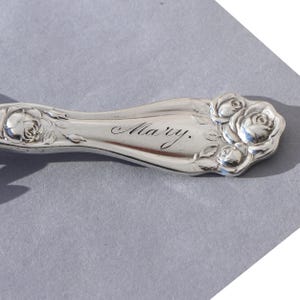 May include: Silverware handle with floral rose design and the engraved name "Mary." The handle is curved and features detailed rose blossoms and leaves. The silverware is likely vintage or antique.