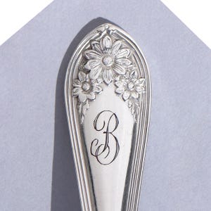 May include: Close-up of a silver spoon with a floral design and the letter "B" engraved on the handle. The spoon has a decorative border and is set against a light gray background. The flowers are detailed and add a touch of elegance.