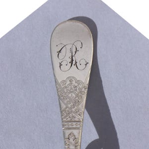 May include: Close-up of a silver spoon with an ornate design. The spoon's bowl features a large, elegant letter "R" in a script font. The handle is decorated with intricate floral and geometric patterns, showcasing a vintage aesthetic.