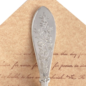 May include: A silver spoon with an ornate floral design. The spoon is resting on a piece of paper with the text "...and to...ume this day for...for poorer, in sick...is health, to love and cherish...To"