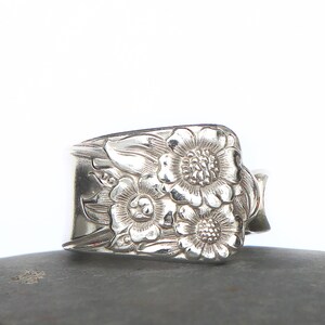 May include: A silver ring with a floral design. The ring features three detailed flowers with intricate petal and center detailing. The ring is made of a shiny, metallic material and is set against a neutral background.