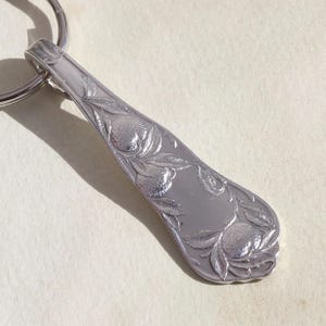 May include: A silver-colored spoon-shaped keychain with an ornate floral and fruit design. The spoon's bowl features detailed carvings of what appear to be oranges and leaves. A silver key ring is attached to the spoon's handle.