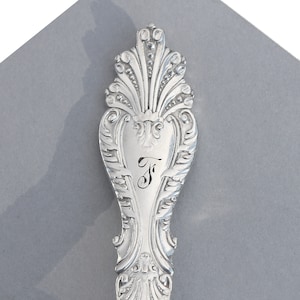 May include: Close-up of a silver spoon handle with an ornate design and the letter "F" engraved on the front.