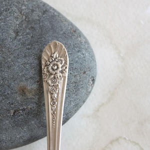 May include: A silver spoon with an ornate floral design on the handle. The spoon is resting on a gray rock.