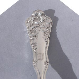 May include: A silver spoon with a floral design on the handle. The handle features a detailed relief of flowers and leaves. The spoon has an engraved letter "I" near the bowl. The spoon is set against a gray background.