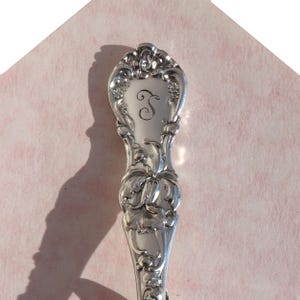 May include: Close-up of the ornate handle of a silver spoon with an engraved letter "S" on the top. The handle is decorated with floral designs.