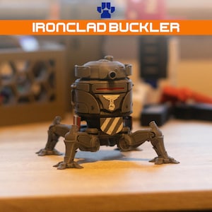 Op de afbeelding: Een grijs, meerbenig robotfiguur met het label IRONCLAD BUCKLER. Het figuur heeft een cilindrisch lichaam met een rode horizontale lijn, een wit embleem aan de voorzijde en diagonale witte en grijze strepen op het onderste gedeelte.