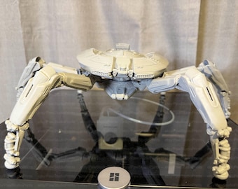 May include: A detailed, light gray model of a mechanical spider with a disc-shaped body and multiple articulated legs. The model has intricate details and appears to be 3D-printed. The legs are positioned to give the impression of movement.