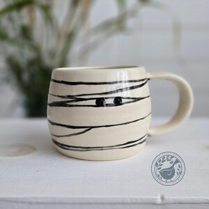 May include: A cream-coloured ceramic mug featuring a hand-painted black line design that wraps around the exterior to resemble mummy bandages, with two small black dots representing eyes visible on the front and a curved handle on the right side.