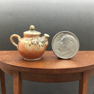 May include: Miniature ceramic teapot with a speckled design in shades of orange, tan, and brown. The teapot is next to a dime for size comparison, sitting on a small wooden table. The teapot has a handle, spout, and lid.
