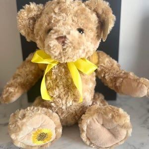 Könnte beinhalten: Ein Teddybär mit hellbraunem Fell und einem leuchtend gelben Band um den Hals. Der Bär hat eine Sonnenblume auf der Unterseite jedes Fußes gestickt. Der Bär sitzt aufrecht.