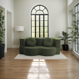 Op de afbeelding: Een moderne, olijfgroene bank met twee ronde, bijpassende kussens. De bank staat op een wit tapijt in een kamer met hardhouten vloeren, een groot boogvenster en potplanten. Een staande lamp is ook zichtbaar.