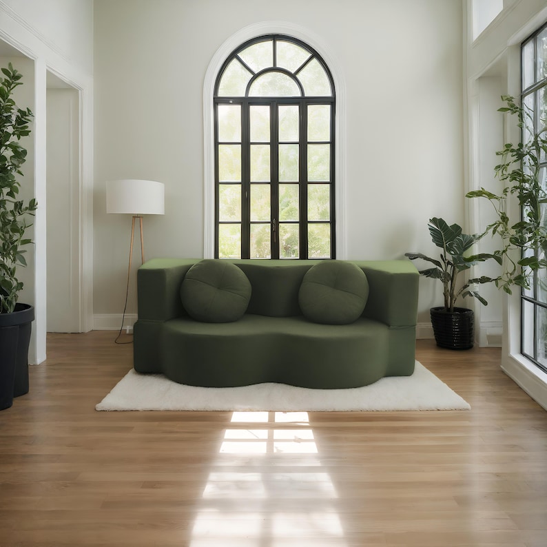 Op de afbeelding: Een moderne, olijfgroene bank met twee ronde, bijpassende kussens. De bank staat op een wit tapijt in een kamer met hardhouten vloeren, een groot boogvenster en potplanten. Een staande lamp is ook zichtbaar.