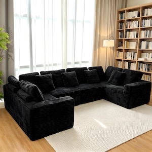 K&ouml;nnte beinhalten: Ein gro&szlig;es, schwarzes Ecksofa aus Cord mit mehreren Kissen. Das Sofa ist U-f&ouml;rmig angeordnet und steht auf einem wei&szlig;en Teppich. Der Raum hat ein gro&szlig;es Fenster mit transparenten Vorh&auml;ngen und ein B&uuml;cherregal voller B&uuml;cher.