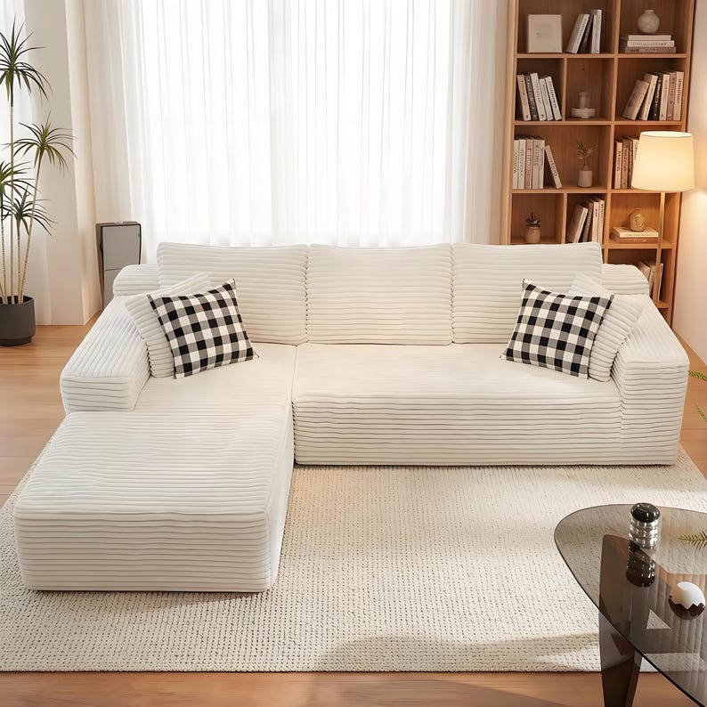 K&ouml;nnte beinhalten: Ein gro&szlig;es, cremefarbenes Ecksofa mit Cordstruktur. Zwei schwarz-wei&szlig; karierte Zierkissen liegen auf dem Sofa. Das Sofa steht auf einem hellen Teppich. Ein B&uuml;cherregal und eine Lampe befinden sich im Hintergrund.