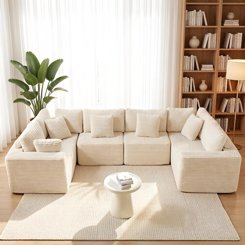 K&ouml;nnte beinhalten: Ein cremefarbenes, modulares Cord-Sofa mit mehreren Kissen, das in U-Form auf einem passenden Teppich angeordnet ist. Vor dem Sofa steht ein kleiner, runder Beistelltisch. Eine gro&szlig;e Zimmerpflanze und ein B&uuml;cherregal befinden sich im Hintergrund.