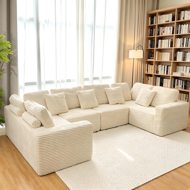 May include: A large, cream-colored sectional sofa with multiple cushions. The sofa has a corduroy texture and is arranged in a U-shape. The room features a large window, a bookshelf, and a white rug.