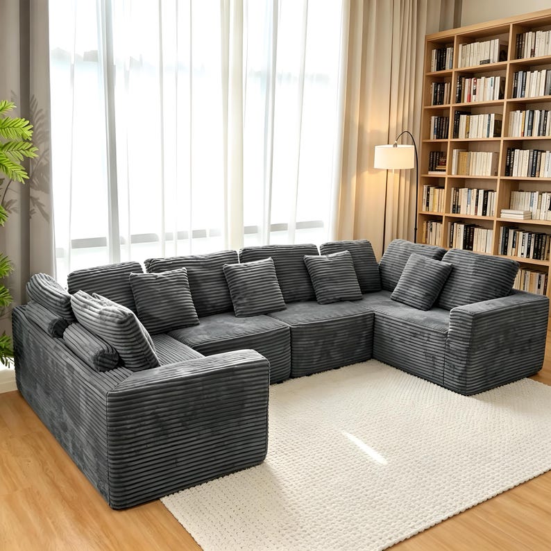 May include: A large, sectional sofa in a U-shape, upholstered in gray corduroy fabric. The sofa is accompanied by several matching throw pillows. The room features a large window with sheer curtains, a bookshelf, and a white rug.