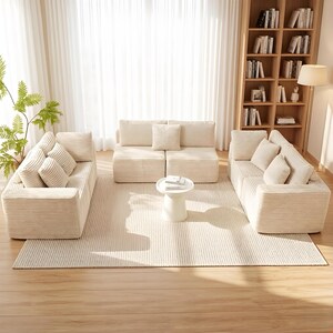 K&ouml;nnte beinhalten: Ein modernes Wohnzimmer-Set mit drei cremefarbenen Cord-Sofas, die um einen kleinen wei&szlig;en Beistelltisch angeordnet sind. Der Raum umfasst einen Teppich in neutralen T&ouml;nen, ein B&uuml;cherregal aus Holz und eine Stehlampe, wodurch eine gem&uuml;tliche Atmosph&auml;re entsteht.