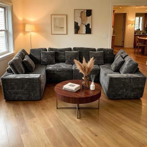May include: A modern living room features a large, sectional sofa in a dark gray corduroy fabric. A round, dark wood coffee table sits in front of the sofa, with decorative items. The room has hardwood floors and neutral-toned walls.
