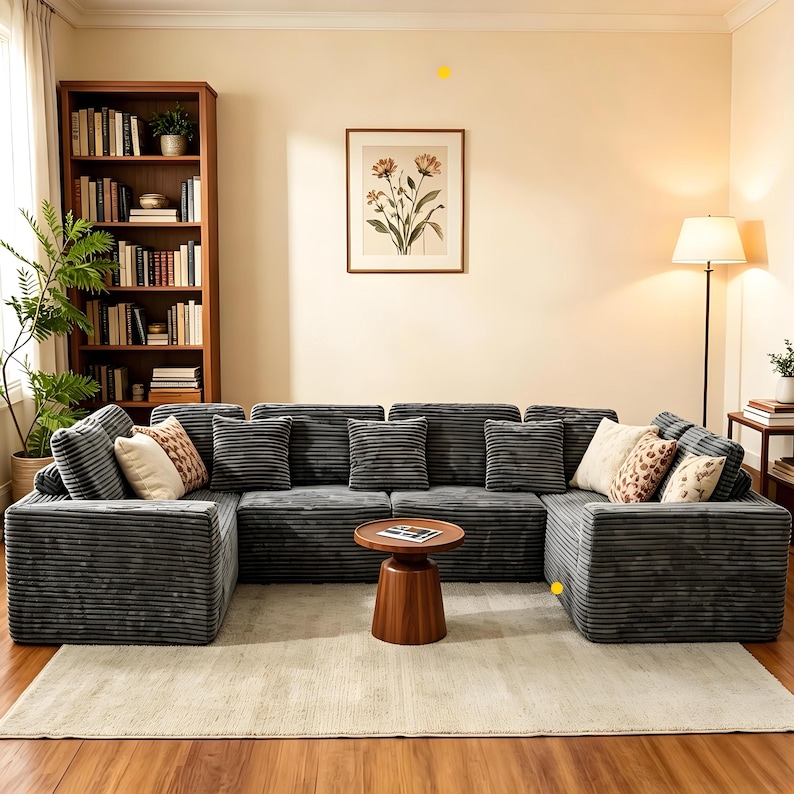 K&ouml;nnte beinhalten: Ein gro&szlig;es, graues Ecksofa aus Cord mit mehreren Kissen und dekorativen Kissen. Vor dem Sofa steht ein kleiner, runder Beistelltisch aus Holz. Der Raum umfasst auch ein B&uuml;cherregal, Kunstwerke und eine Stehlampe.