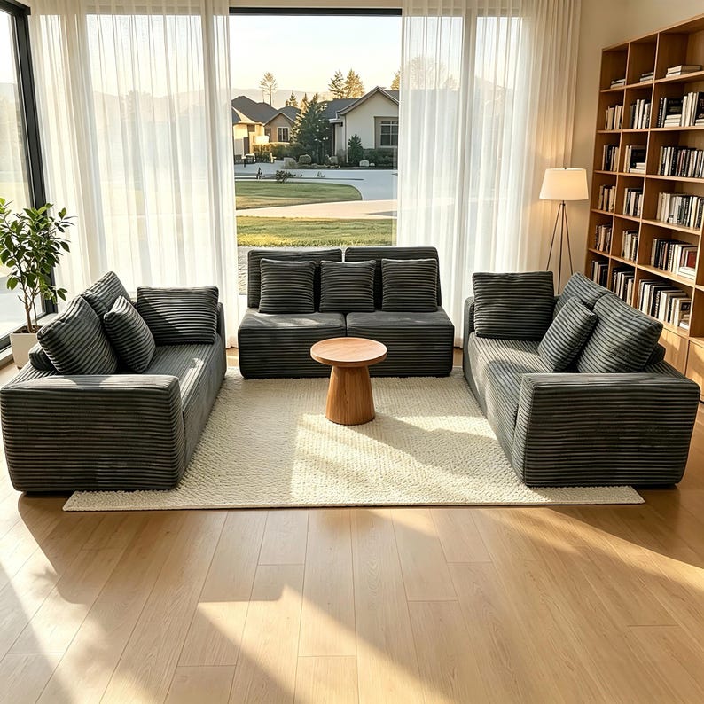 May include: A modern living room set featuring a gray corduroy sofa, loveseat, and armchair with matching throw pillows. A small, round wooden coffee table sits on a cream-colored rug. The room has large windows and a bookshelf.