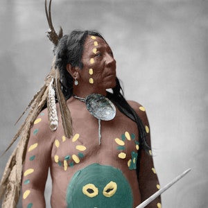 May include: A Native American man wearing traditional clothing and paint. He is holding a spear and a shield with a painted image of a rider on a horse. The shield is decorated with a yellow, green, and brown design.