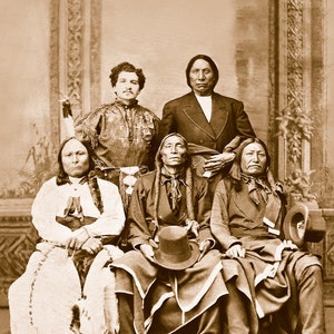 May include: A black and white photograph of five men, four of whom are Native American, sitting in a row. The men are wearing traditional clothing and hats. The photo is likely from the late 1800s or early 1900s.