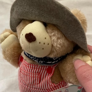 May include: A plush teddy bear wearing a brown hat, red and white striped overalls, and a blue bandana. The bear has a beige snout and is wearing tan shoes. A Wendy's tag is attached.