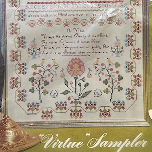 May include: A framed cross-stitch sampler kit titled "Virtue Sampler" by Paragon Needlecraft. The sampler features an alphabet, floral motifs, and a verse. The kit is designed to fit a 17x23 inch frame and includes stamped linen and embroidery thread.