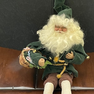 May include: A green and brown elf doll with a white beard and a basket in its hand. The elf is sitting on a brown surface.