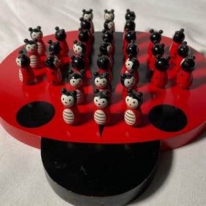 May include: A wooden ladybug-shaped game board with a red base and black accents. The board features numerous small, wooden ladybug game pieces. The game pieces are painted with black heads and red bodies, and are arranged in rows.