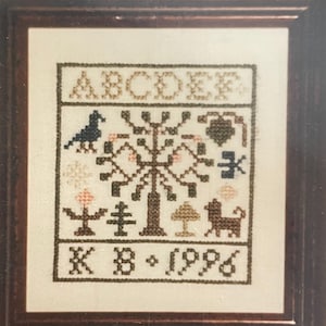 May include: A framed cross-stitch embroidery featuring a stylized tree with various animals and plants. The letters "ABCDEF" are stitched at the top, and the date "K B - 1996" is stitched at the bottom.