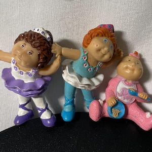 May include: Three vintage Cabbage Patch Kids figurines. One is a ballerina in a purple tutu, another is in a blue outfit, and the third is a baby in pink pajamas. The figurines are made of plastic and are in good condition.