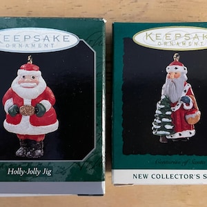 May include: Two Christmas ornaments in their original boxes. One ornament is a Santa Claus figure in a red suit, holding a gold belt buckle. The other is a Santa Claus figure holding a Christmas tree. The boxes are green and have the text "Keepsake Ornament".